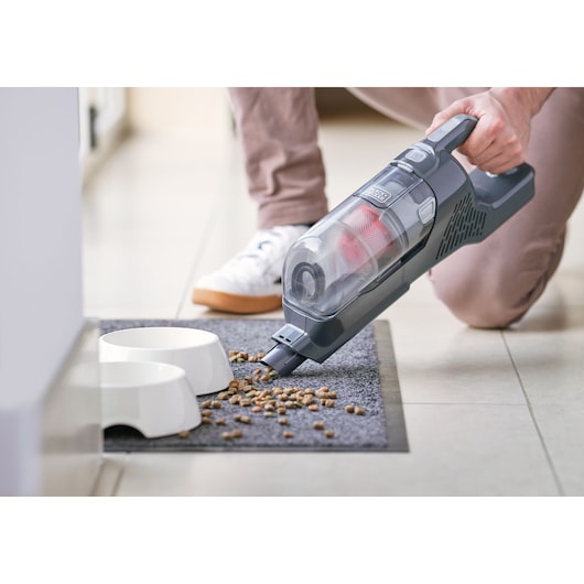 A person using a BLACK+DECKER handheld vacuum cleaner to pick up scattered pet food from a carpet near two white pet bowls.