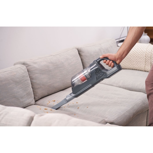 A person is using a BLACK+DECKER handheld vacuum cleaner to clean crumbs from a light-colored sofa.