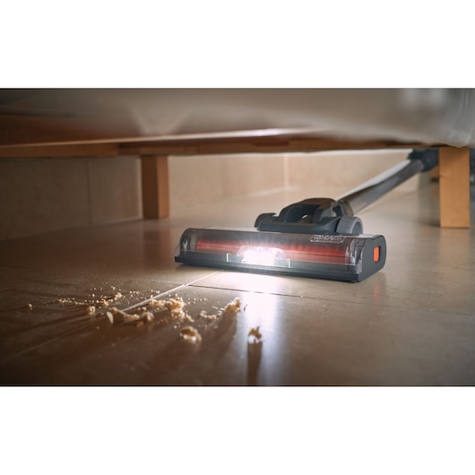 A BLACK+DECKER cordless vacuum cleaner with an LED light is being used to clean crumbs from a tiled floor underneath a piece of furniture.