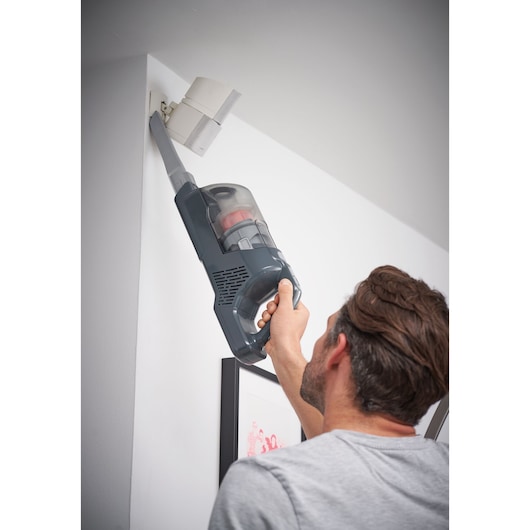 A person using a BLACK+DECKER cordless vacuum cleaner with a long nozzle attachment to clean dust from a high corner near a wall-mounted fixture.