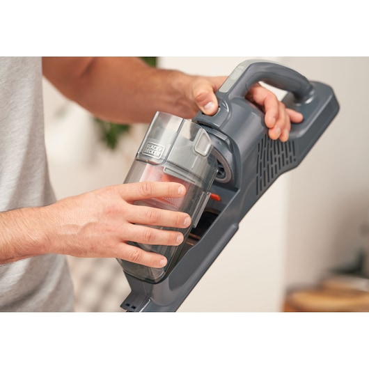 A close-up of a person holding and removing the dust container from a BLACK+DECKER cordless vacuum cleaner.
