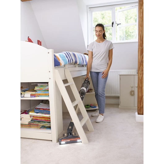A person is vacuuming the carpeted floor in a brightly lit children's bedroom using a BLACK+DECKER cordless vacuum cleaner. The room features a white bunk bed with bookshelves, colorful bedding, and a window.
