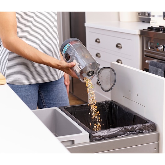 A person empties the dust and debris from the canister of a BLACK+DECKER vacuum cleaner into a kitchen trash bin lined with a black plastic bag.
