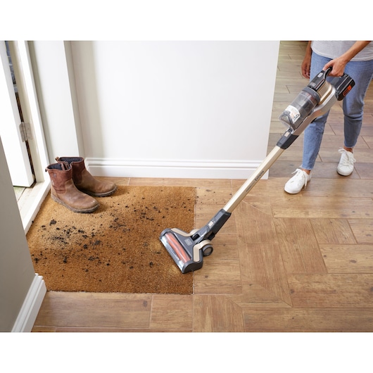 A person is using a BLACK+DECKER cordless stick vacuum cleaner (model BHFEV36B2DW) to clean dirt from a brown doormat next to a pair of muddy boots by the entrance on a wooden floor.