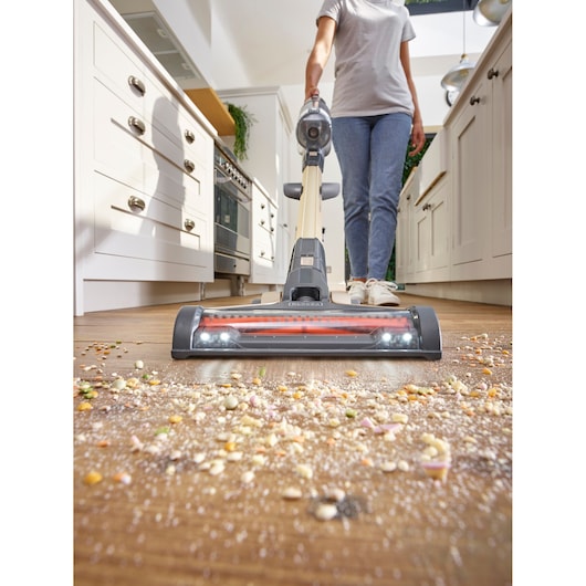 A person using a BLACK+DECKER cordless vacuum cleaner to clean scattered crumbs and debris from a hardwood kitchen floor.