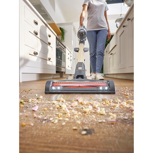 A person is vacuuming a wooden kitchen floor with a BLACK+DECKER cordless stick vacuum, picking up scattered crumbs and debris. The scene is in a bright modern kitchen.