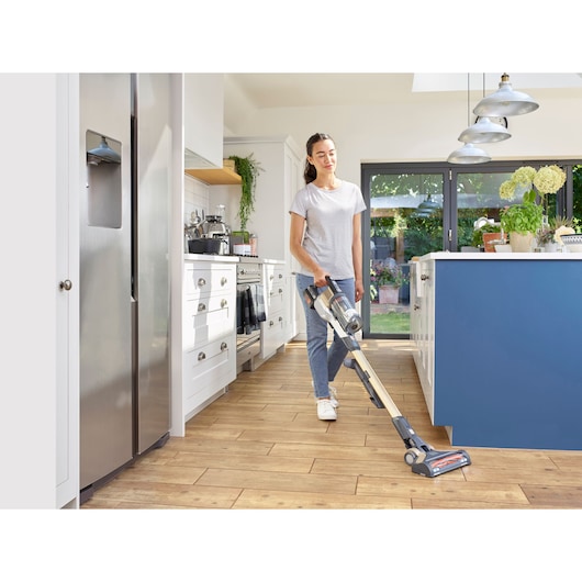 A person using a BLACK+DECKER cordless stick vacuum cleaner (model BHFEV36B2DW) to clean a modern kitchen with wooden floors. The kitchen is bright with white cupboards, a blue island, stainless steel refrigerator, and large glass doors leading outside. The person's face is blurred for privacy.