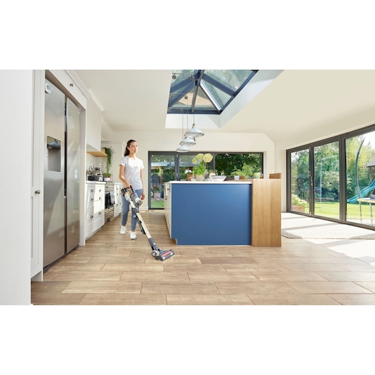 A person using a BLACK+DECKER cordless vacuum cleaner in a modern kitchen with wooden floors, blue island, large windows, and skylight, with a view of a green garden outside.
