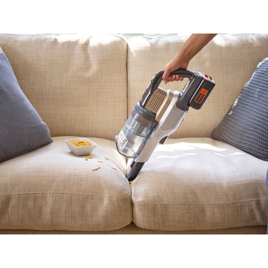 A person using a BLACK+DECKER cordless vacuum cleaner to clean crumbs from a beige sofa, with a bowl of snacks next to the crumbs.