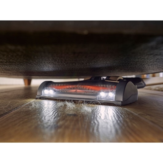 Close-up view of a vacuum cleaner head with LED lights, cleaning dust and pet hair under a piece of furniture on a wooden floor.