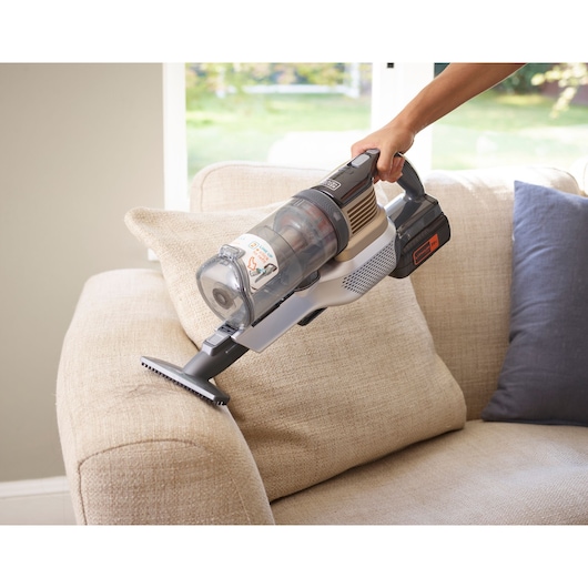 A person is using a BLACK+DECKER cordless handheld vacuum cleaner to clean a beige fabric sofa in a living room.