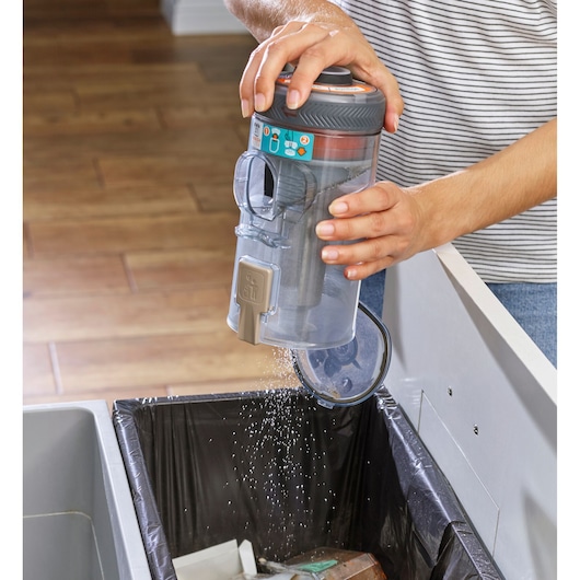 A person is emptying the dust container of a BLACK+DECKER cordless vacuum cleaner over a trash bin, letting dust and debris fall out.