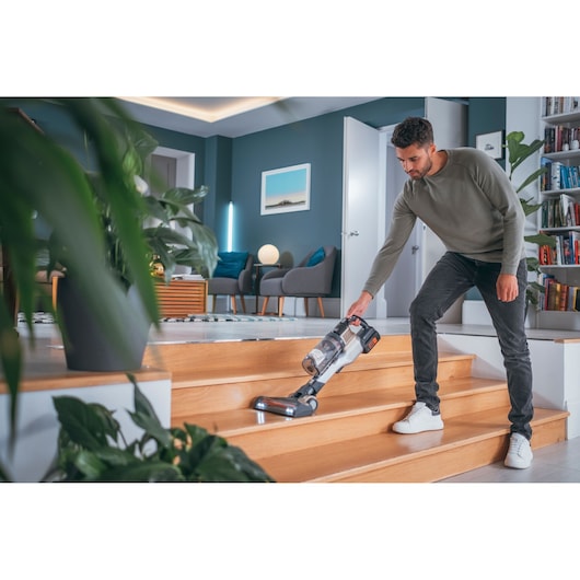 A person cleaning wooden stairs in a modern living room with a BLACK+DECKER cordless vacuum cleaner. The vacuum has LED lights and is designed for easy maneuverability on stairs.