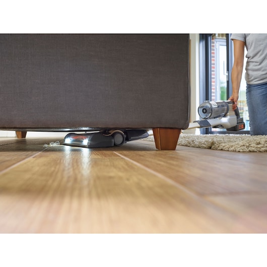 A cordless vacuum cleaner being used to clean underneath a sofa on a wooden floor, with a person holding the handle.