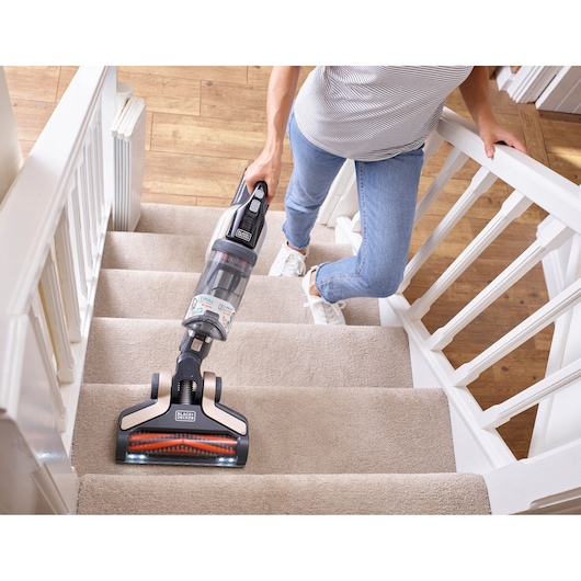 A person cleaning carpeted stairs with a BLACK+DECKER cordless vacuum cleaner. The vacuum has LED lights on the brush head and a transparent dust container.