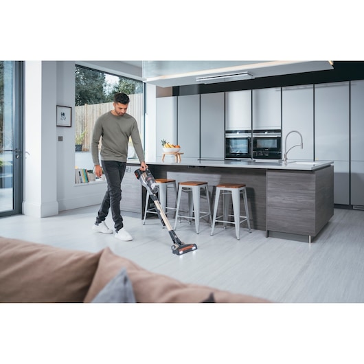 A person is using a BLACK+DECKER cordless vacuum cleaner to clean the floor in a modern, bright kitchen with an island and stools. The vacuum is lightweight and easy to maneuver.