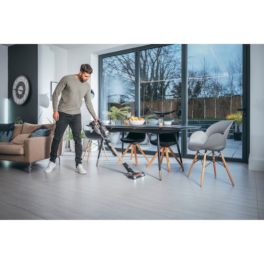 A person is vacuuming the floor of a modern living and dining area using a BLACK+DECKER cordless stick vacuum cleaner. The room features large windows, contemporary furniture, and a dining table with chairs.