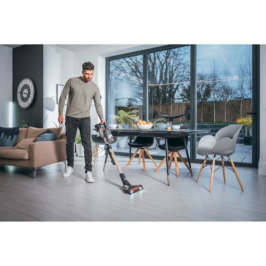 A person using a BLACK+DECKER cordless stick vacuum cleaner to clean the floor in a modern living and dining area with large windows and contemporary furniture.