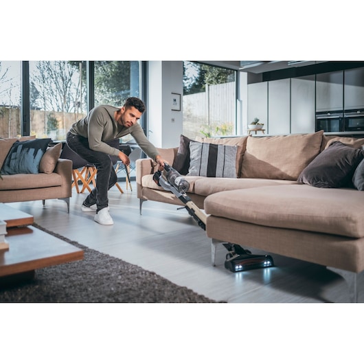 A person using a BLACK+DECKER cordless vacuum cleaner to clean under a beige sofa in a modern living room with large windows.