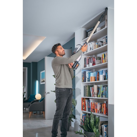 A person is using a BLACK+DECKER cordless vacuum cleaner with an extended nozzle to clean the top corner of a white bookshelf in a modern living room.