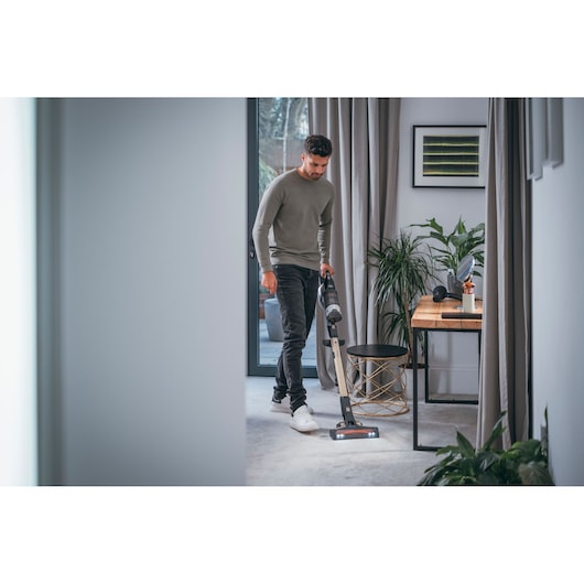 A person vacuuming a carpet in a modern living space using a BLACK+DECKER cordless vacuum cleaner. The room features a desk, a chair, indoor plants, curtains, and wall art.