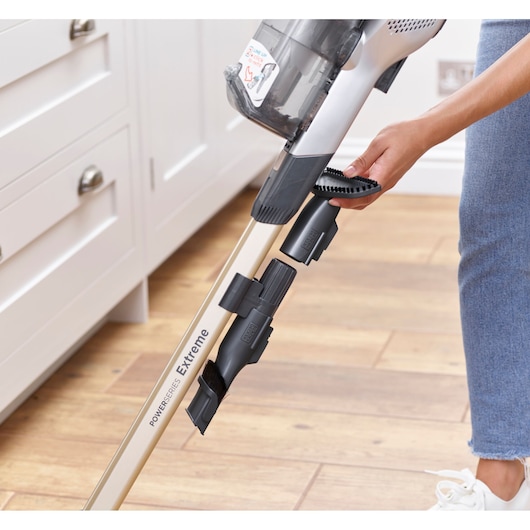 A person attaches a cleaning accessory to a BLACK+DECKER PowerSeries Extreme cordless vacuum cleaner in a kitchen with wooden flooring.