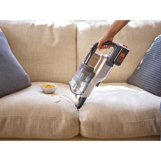 A person using a BLACK+DECKER cordless vacuum cleaner to clean crumbs from a beige sofa, with a bowl of snacks next to the crumbs.