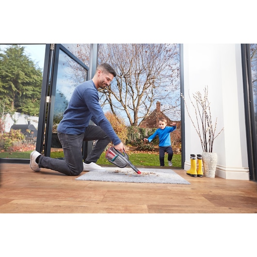 BLACKDECKER BHHV315B handheld vacuum cleaning rug near open door with garden view, boots and vase visible.