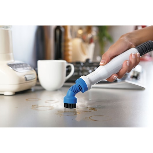 A person is using a handheld cleaning device with a blue brush attachment to clean stained rings from a kitchen countertop. In the background, there is a white mug, a blender, and a stove.