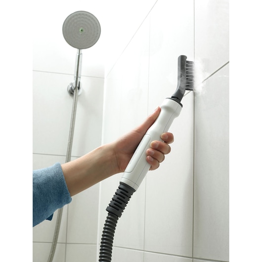 A steam cleaning device is being used to clean grout lines between shower tiles. The hand-held tool emits steam onto the tile joints, helping remove dirt and residue.