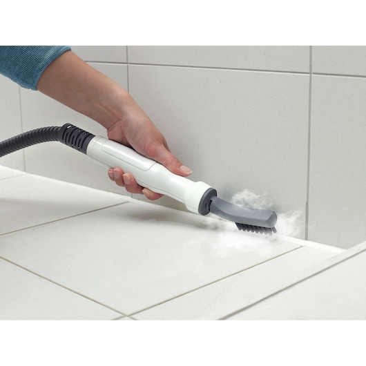 A hand is holding a BLACK+DECKER steam cleaner with a brush attachment, cleaning the grout between white tiled surfaces, with visible steam coming out.