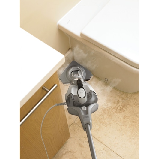 A BLACK+DECKER steam mop cleaning a tiled floor next to a bathroom cabinet and toilet, with visible steam coming from the mop head.