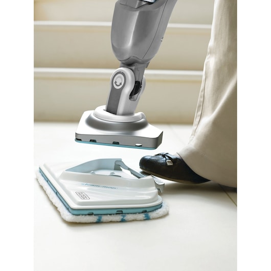 A close-up of a BLACK+DECKER steam mop being attached to its cleaning pad, with a person pressing the pad into place using their foot. The scene shows the mop and pad on a light tiled floor.