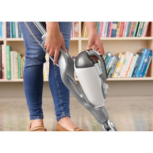 A person using a BLACK+DECKER BHSM168D steam mop on a hard floor, with a bookshelf filled with books in the background.