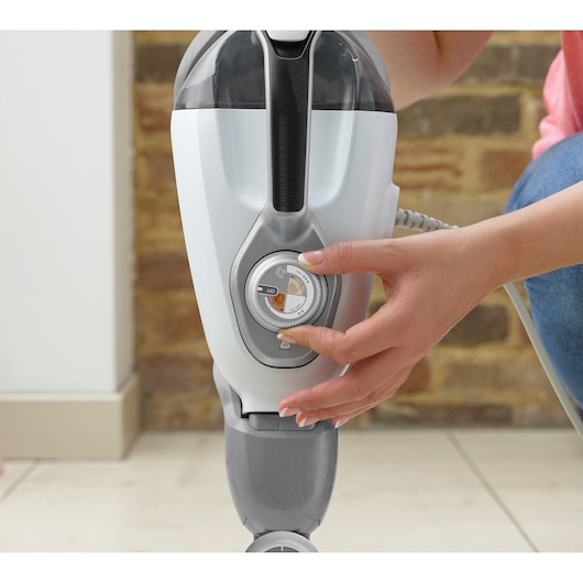 Close-up of a person adjusting a control dial on a BLACK+DECKER cleaning appliance, with a brick wall and tiled floor in the background.