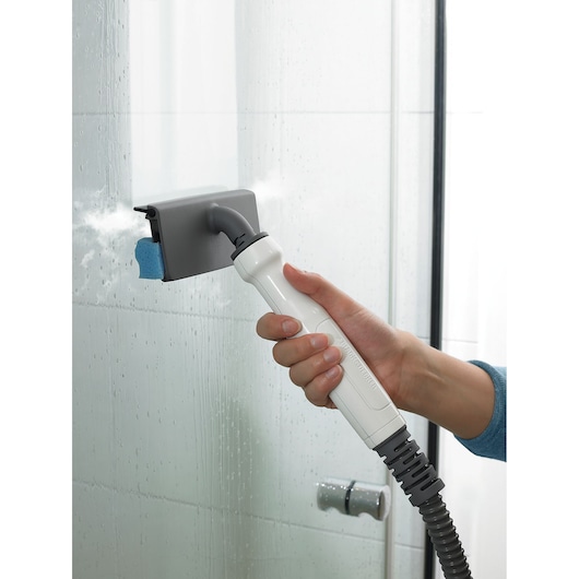 A person using a BLACK+DECKER handheld steam cleaning device to clean a glass shower door with visible steam and water droplets.