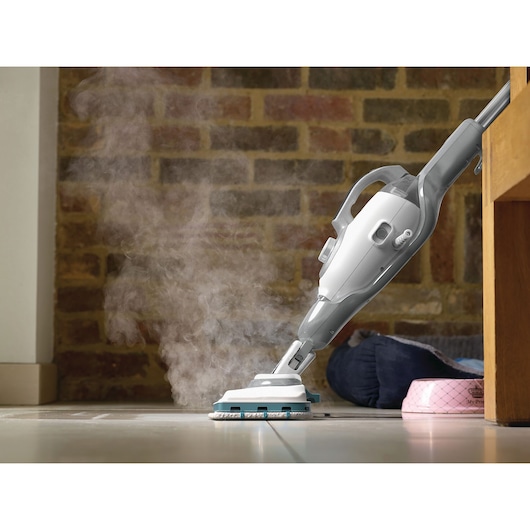 A steam mop cleaning a tiled floor with visible steam, next to a brick wall and some pet accessories.
