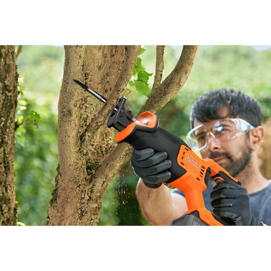 BLACK+DECKER BLCR78M1 reciprocating saw cutting a tree branch outdoors, person wearing gloves and safety glasses.