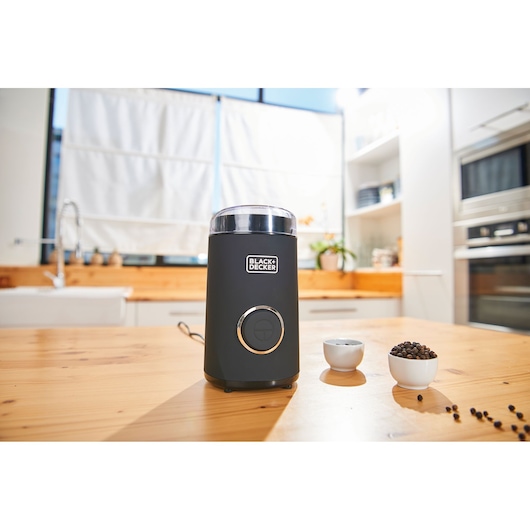 BLACKDECKER BXCG150E coffee grinder in black with transparent lid on kitchen table, bowls of coffee beans nearby.