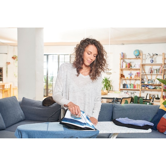 BLACKDECKER BXIR2400E steam iron in use on ironing board, modern living room setting, shirts folded nearby.