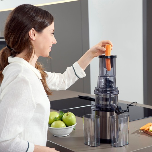 BLACKDECKER BXJE200E juicer in use, carrot being inserted, with two clear containers and apples nearby.
