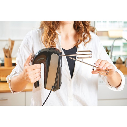 Person attaching metal beaters to BLACKDECKER BXMX500E hand mixer in kitchen setting.