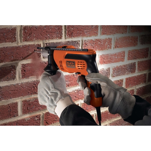 A person wearing protective gloves uses an orange and black BLACK+DECKER electric drill to work on a red brick wall.