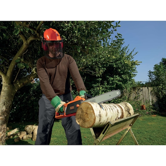 A person wearing protective gear uses a BLACK+DECKER electric chainsaw to cut a log supported on a stand in a garden.