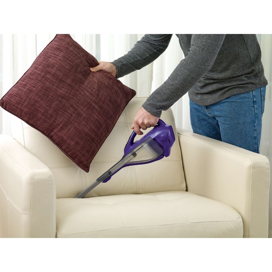 BLACKDECKER DVA325JP07 handheld vacuum cleaning a beige armchair with a person lifting a red cushion.