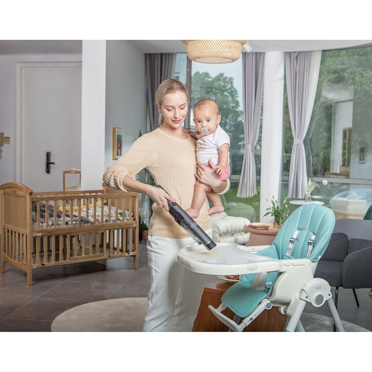 A person holding a baby and using a BLACK+DECKER handheld vacuum cleaner to clean the surface of a high chair in a home setting. A crib is visible in the background, and the space appears to be tidy and well-lit.