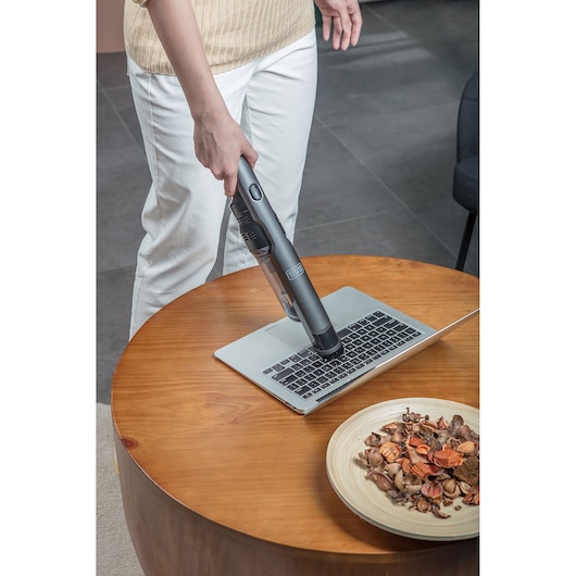A person uses a BLACK+DECKER handheld vacuum cleaner to clean the keyboard of a laptop computer on a wooden table. A plate with dried potpourri is also on the table.