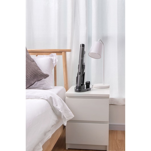 A BLACK+DECKER handheld vacuum cleaner sits on a charging base on a white bedside table next to a bed with white linens and a gray pillow. There is also a white desk lamp on the table, and light curtains in the background.