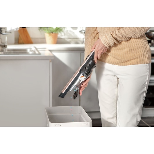 A person empties a handheld vacuum cleaner into a trash bin in a kitchen setting.