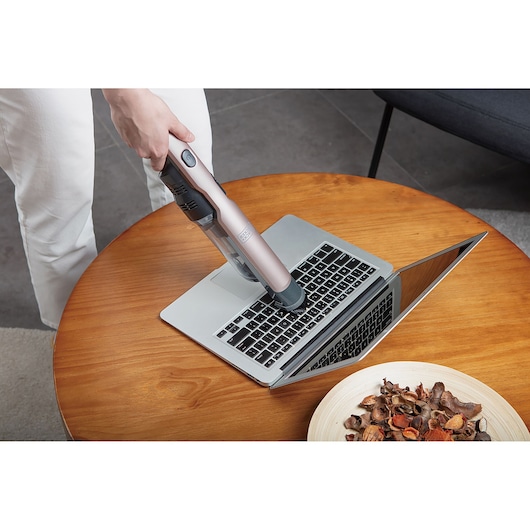 A person uses a BLACK+DECKER handheld vacuum cleaner to clean the keyboard of an open laptop on a wooden table. There is a bowl of dried flower petals on the table.